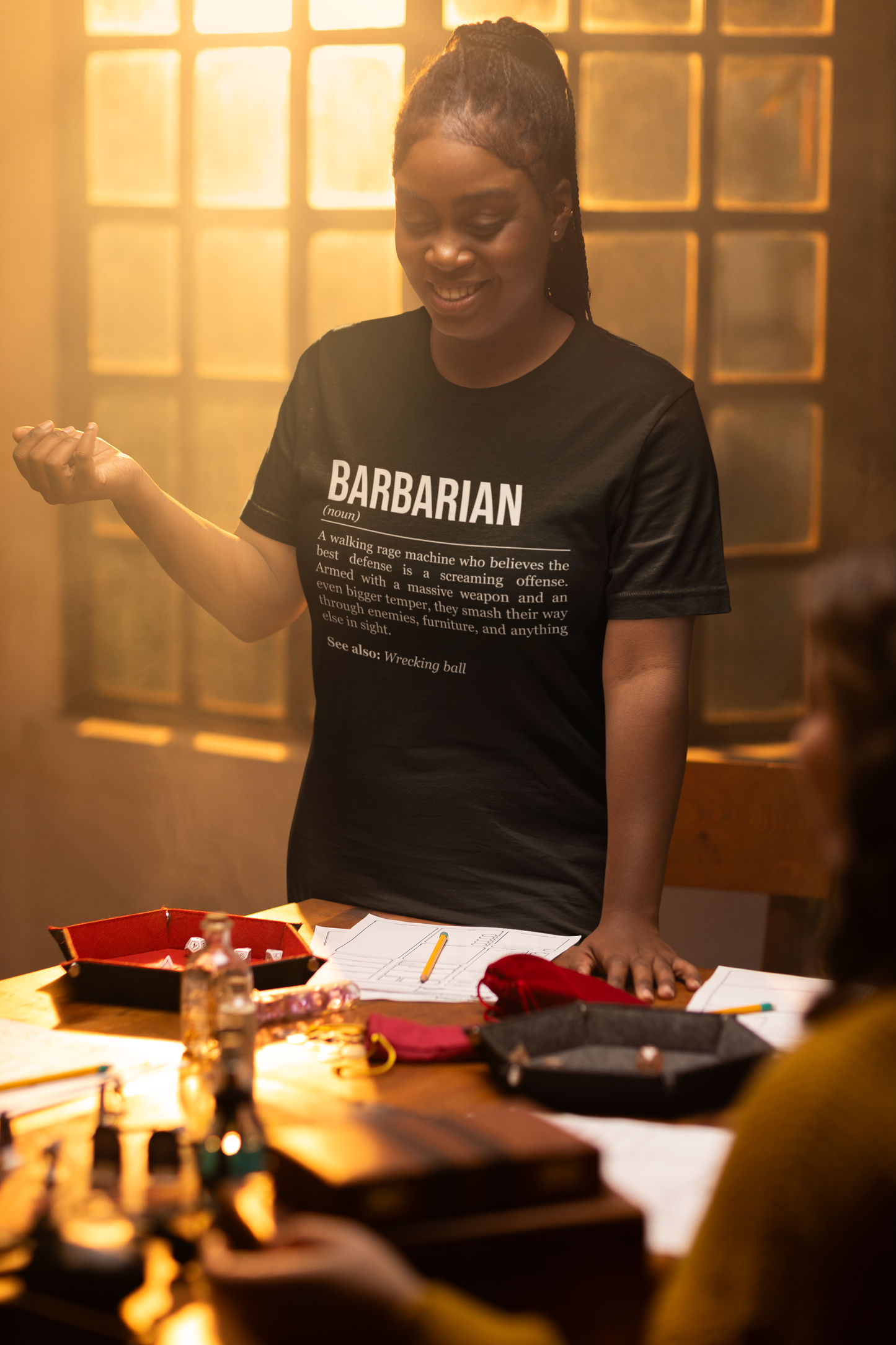 A standing woman playing an RPG at a table, wearing a black barbarian T-shirt showing the definition of the class Barbarian.