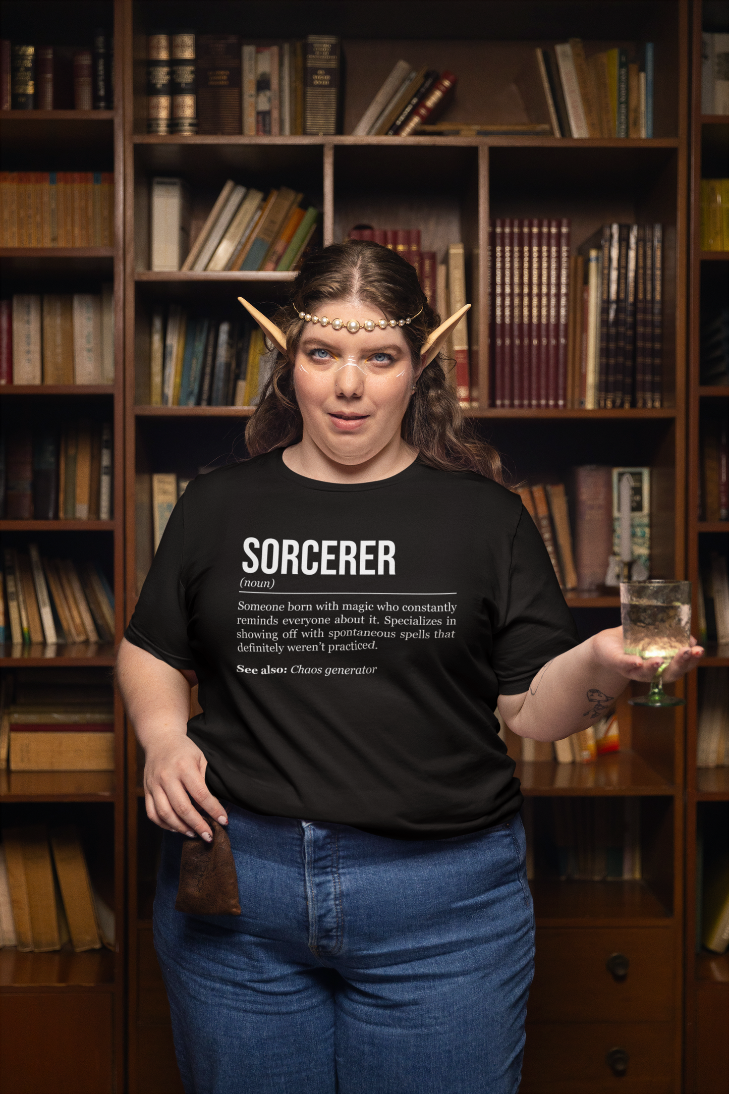 A standing woman with elf ears holding a pouch and a glass of water, wearing a black sorcerer T-shirt showing the definition of the class Sorcerer.