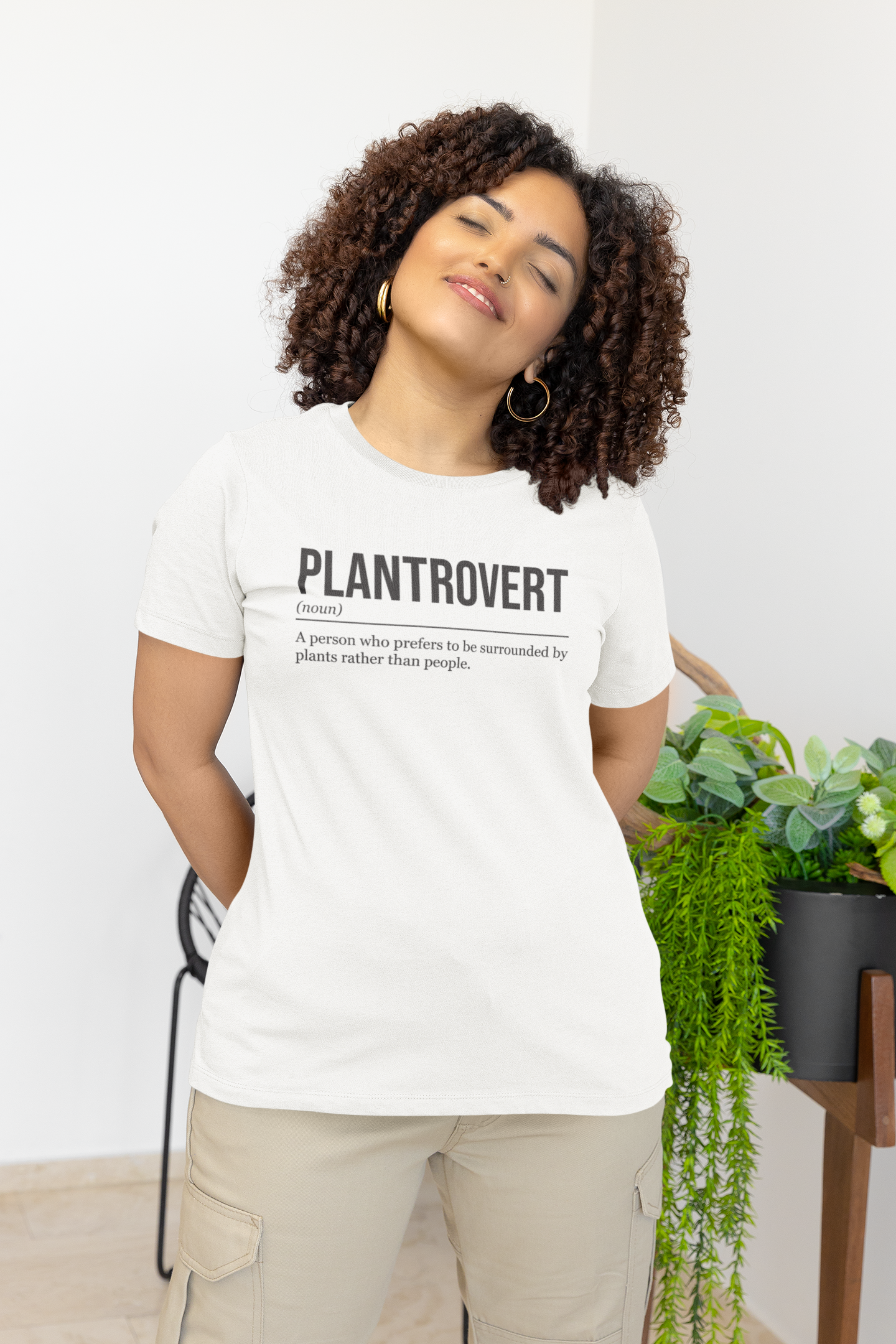 A standing woman with closed eyes and her arms behind her, front view, wearing a white plant T-shirt showing the definition of the word Plantrovert.