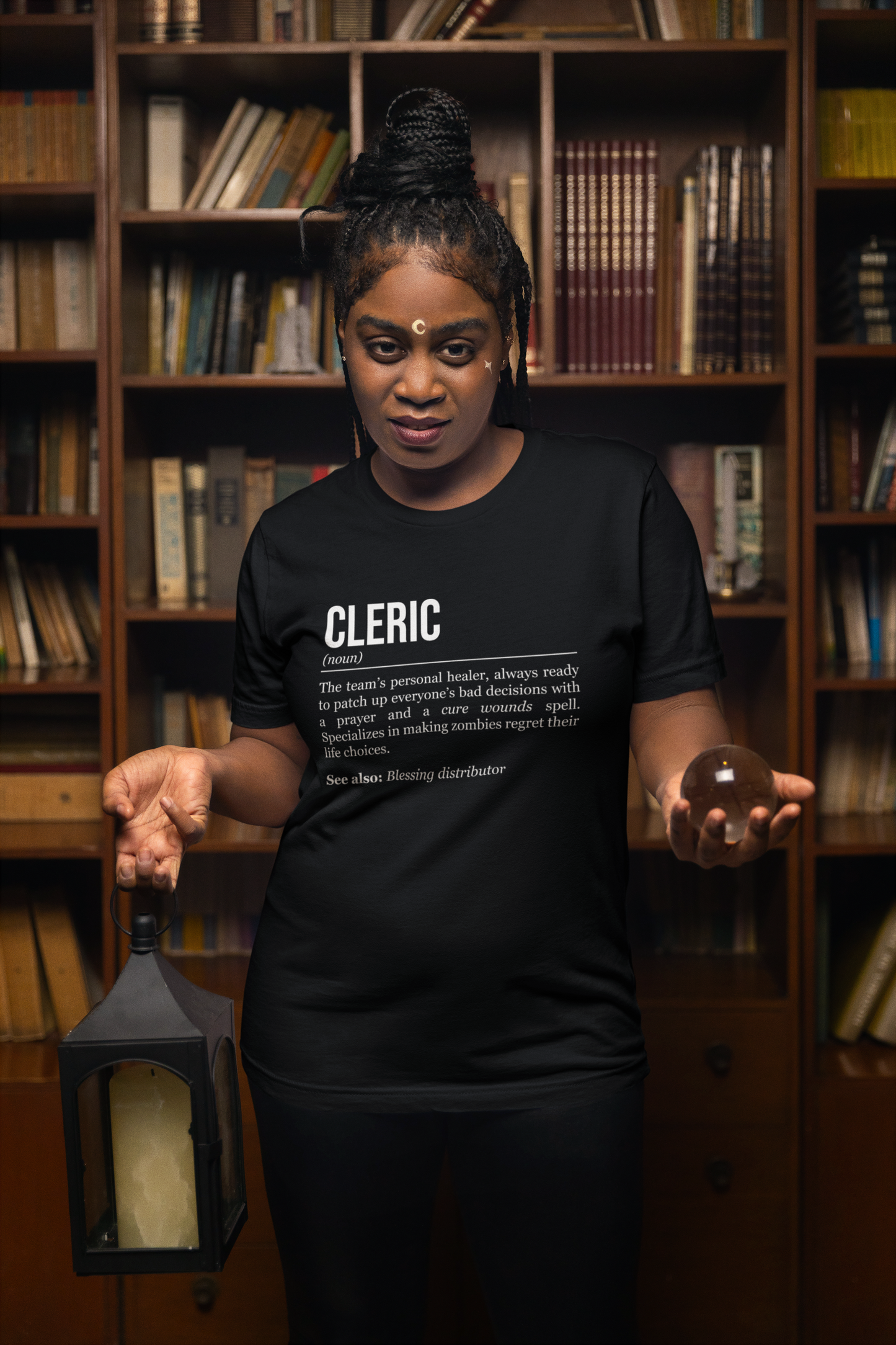A standing woman holding a lantern and a glass ball, wearing a black cleric T-shirt showing the definition of the class Cleric. 