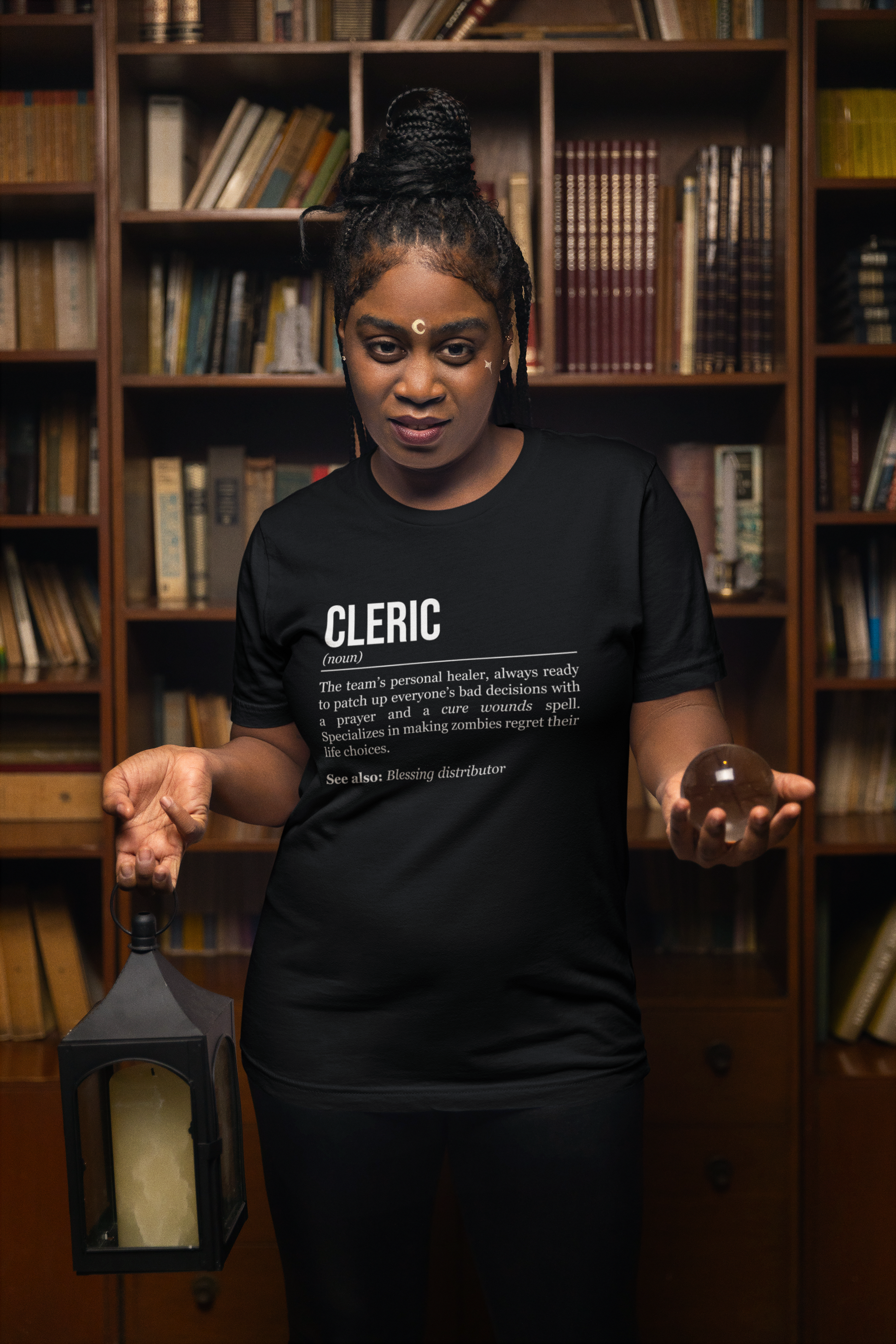 A standing woman holding a lantern and a glass ball, wearing a black cleric T-shirt showing the definition of the class Cleric. 