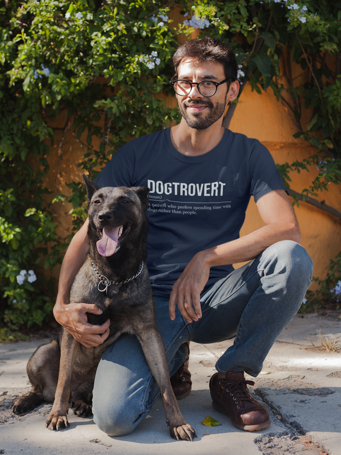 A man kneeling and hugging a dog by his side. The man is wearing a navy blue dog T-shirt showing the definition of the word Dogtrovert.