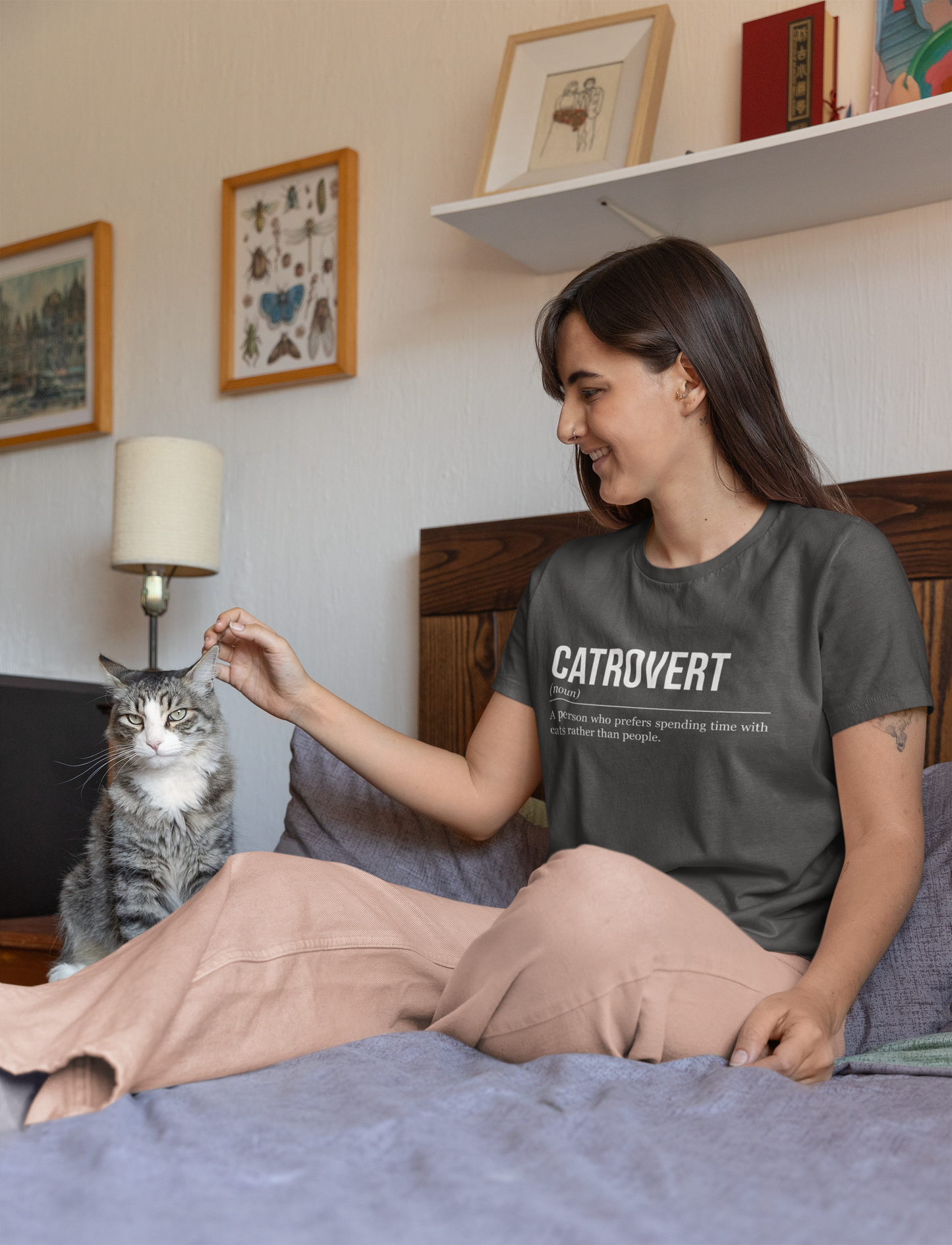 A woman sitting on a bed next to a grey cat. The woman is petting the cat and wearing  a grey plant T-shirt showing the definition of the word Catrovert.