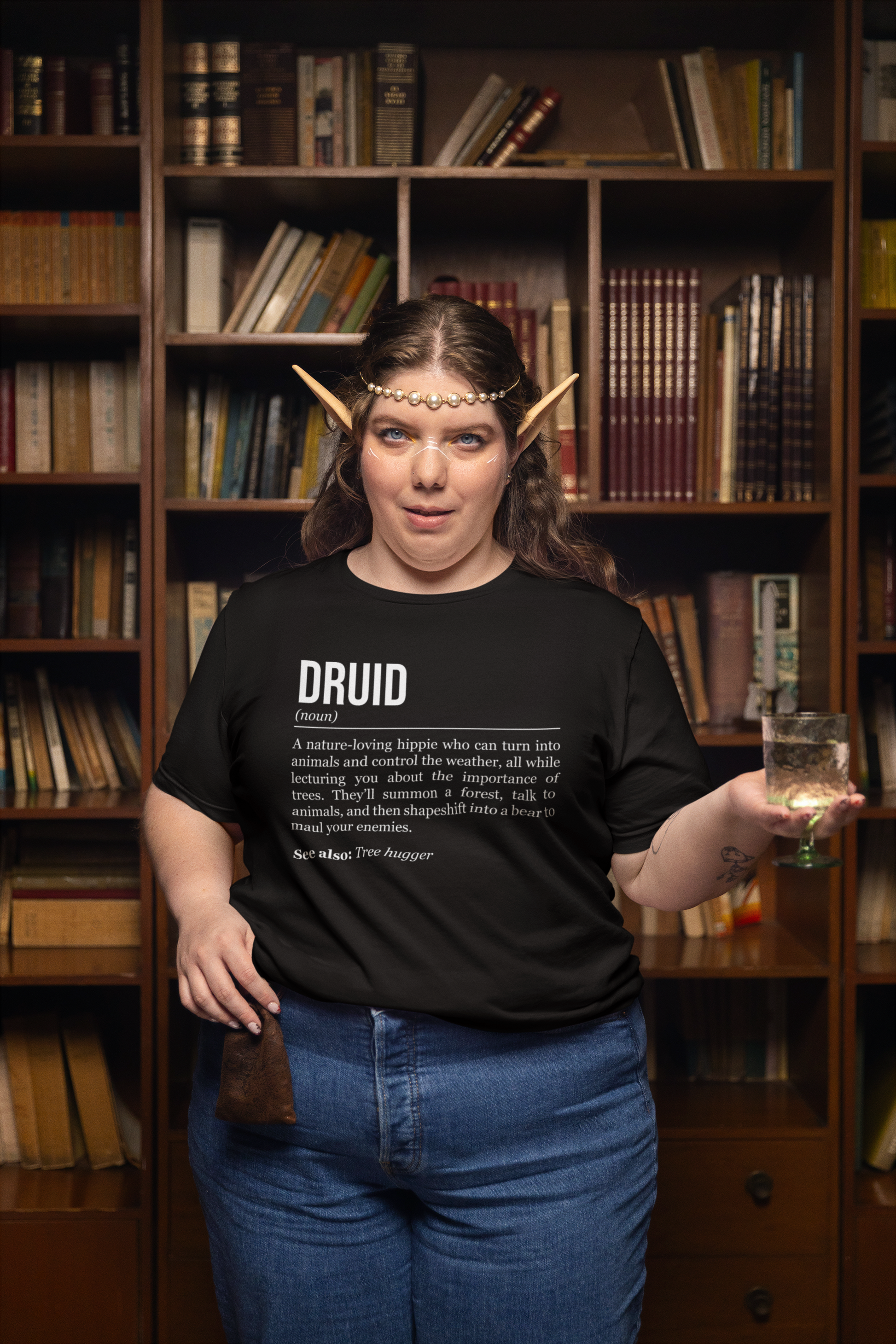 A standing woman with elf ears holding a pouch and a glass of water, wearing a black druid T-shirt showing the definition of the class Druid. 